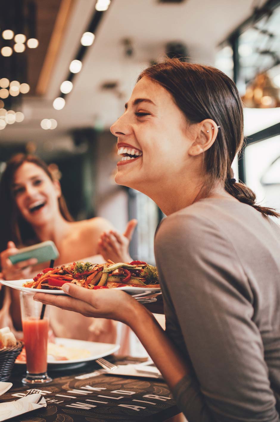 Woman laughing while holding a plate of grilled vegetables at a restaurant table with friends nearby